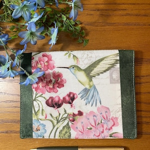 May include: A green fabric covered journal with a hummingbird and floral print. The journal has a green fabric border and a gold pen is lying next to it.