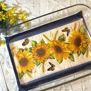 May include: A blue and white quilted hot pad with a sunflower and butterfly design. The hot pad is in a glass baking dish.