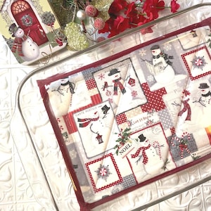 May include: A red and white quilted hot pad with a snowman theme. The hot pad features a variety of snowmen, snowflakes, and other winter-themed designs. The hot pad is in a glass baking dish.