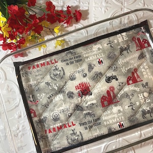 May include: A gray and white quilted hot pad with a black border. The pad features a repeated pattern of farm-themed imagery, including tractors, barns, and the text "FARMALL".