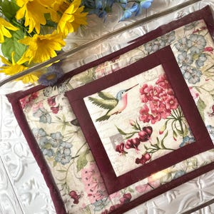 May include: A quilted hot pad with a floral design and a hummingbird in the center. The hot pad has a maroon border and is made of fabric with a white background.