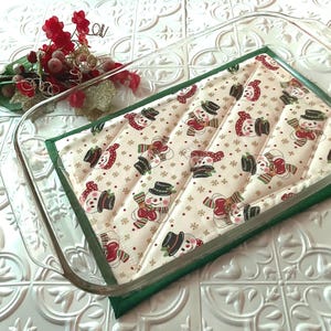 May include: A green and white quilted hot pad with a snowman pattern. The hot pad is sitting on top of a glass baking dish.