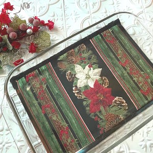 May include: A black and green fabric pot holder with a Christmas theme featuring pine cones, holly berries, and poinsettia flowers. The pot holder is in a clear glass baking dish.