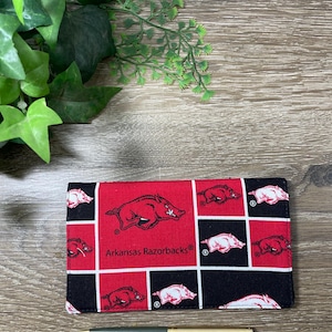 May include: A rectangular fabric checkbook cover featuring a repeating pattern of red and black squares. The design includes white and red Arkansas Razorbacks hog logos and the text Arkansas Razorbacks. A gold and dark green pen lies below.