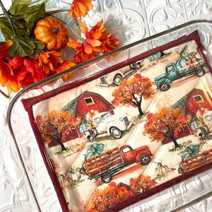 May include: A red and white quilted hot pad with a fall theme. The pad features a red barn, a white truck, a red truck, and pumpkins. The pad is in a glass baking dish.