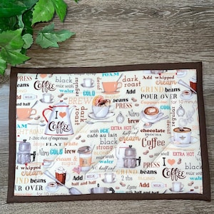 May include: A rectangular, quilted coffee-themed placemat with a brown border. The cream-colored center features coffee-related words and phrases, along with coffee cup illustrations and a red heart with the words "I Coffee".