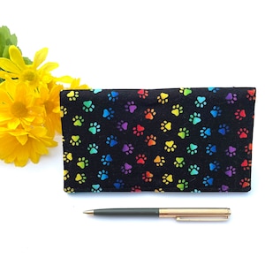 May include: A black fabric wallet with a rainbow paw print pattern. The wallet is lying flat on a white surface with a pen next to it.