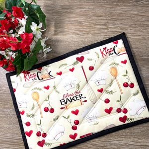 May include: Cream-colored quilted hot pad with a black border. The pad features a repeating pattern of red cherries, hearts, chef hats, and kitchen utensils. Text includes "Kiss the Cook" and "Bless the Baker."