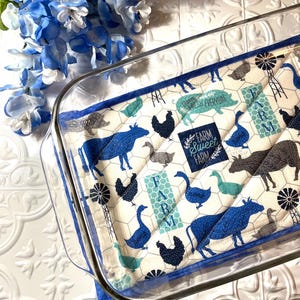 May include: A blue and white quilted hot pad with a farm theme. The pad features cows, chickens, geese, and a windmill. The text "Farm Sweet Farm" is printed on the pad.