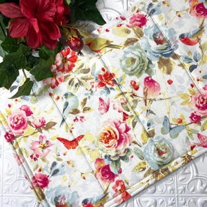May include: A floral-patterned placemat featuring roses, birds, and butterflies in shades of pink, blue, and yellow on a white background. The placemat has a quilted appearance and a gold border. Decorative red flowers and greenery are in the background.