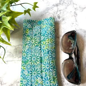 May include: A blue and green floral patterned fabric eyeglass case with a snap closure. The case is lying on a white marble surface next to a pair of sunglasses.