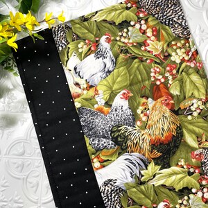 May include: A fabric item featuring a colorful chicken and foliage print. The design includes white chickens with red combs, set against a backdrop of green leaves and red berries. A black border with white polka dots is on the left side.