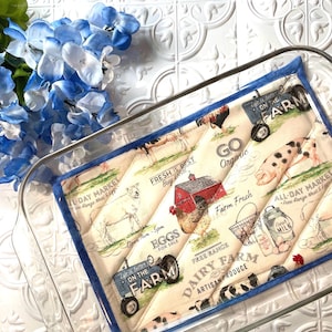 May include: A quilted kitchen hot pad with a blue trim. The fabric features a farmhouse theme with images of farm animals, a red barn, and text that says "Life is better on the farm", "Fresh is best", "Go Organic", "Free Range Dairy Farm", and "All-Day Market".