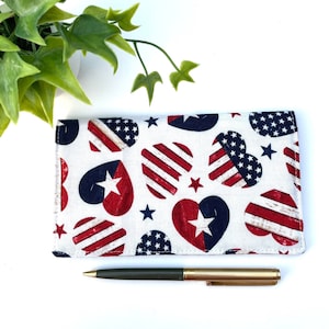 May include: A white fabric checkbook cover with a red, white, and blue heart pattern. The hearts have a distressed wood look and feature stars and stripes. A pen is lying on top of the cover.