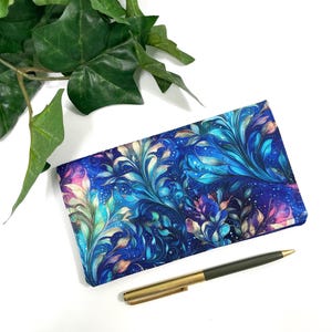 May include: A rectangular checkbook cover with a vibrant, swirling floral pattern in shades of blue, teal, and pink. A gold-toned pen rests beside the cover. Green leafy vines are in the background.