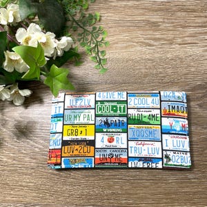 May include: A rectangular fabric item featuring a collage of colorful license plates from various U.S. states. The plates display a variety of colors, text, and designs, creating a vibrant and eye-catching pattern. The item is placed on a wooden surface.