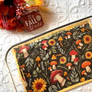 May include: A quilted hot pad with a dark green background and a pattern of red mushrooms, yellow sunflowers, and green leaves. The hot pad has a yellow border and is sitting in a clear glass baking dish. The text "It's Fall y'all" is visible in the background.