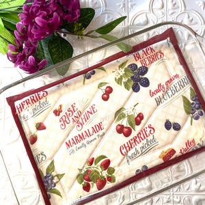 May include: A red and white quilted kitchen hot pad with a patchwork design featuring various berries, cherries, and the text "Rise and Shine Marmalade with Locally Grown Fruit".