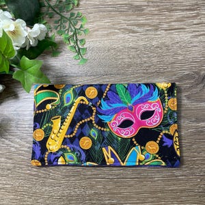 May include: A rectangular wallet with a black background, featuring a colorful Mardi Gras mask, gold coins, and a saxophone. The mask is pink, blue, and purple. The wallet has a patterned design with peacock feathers.