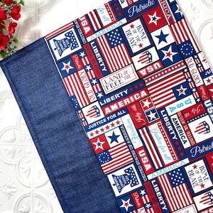 May include: A patriotic-themed fabric item featuring a blue denim border and a central pattern of American flags, stars, and patriotic text. The design incorporates red, white, and blue colors, with phrases like "Liberty" and "USA".