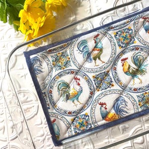 May include: A white quilted hot pad with a blue border features a repeating pattern of colorful roosters in blue, yellow, and red surrounded by blue and white floral designs.