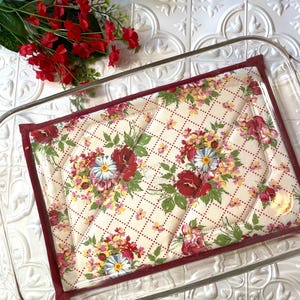 May include: A quilted floral baking dish liner with a red border. The liner is placed inside a clear glass baking dish. The liner has a white background with a red and white diamond pattern and a floral print.