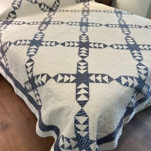 May include: A white bed quilt featuring a repeating navy blue geometric pattern composed of small triangles and dots. The quilt has a grid-like structure with cross-shaped motifs connecting large white squares, finished with a matching patterned border.