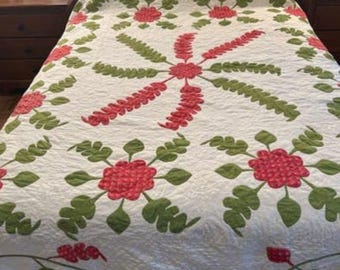 May include: A white quilt with a floral pattern. The quilt features red and green flowers with leaves. The quilt is on a bed with a wooden headboard.