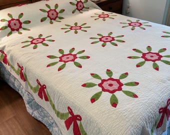 May include: A white quilt with a floral pattern featuring pink and red flowers with green leaves. The quilt has a green vine and red bow border. The quilt is on a bed with a wooden headboard.
