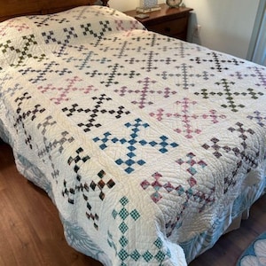 May include: A white quilt with a patchwork design featuring various shades of blue, green, pink, and brown. The quilt is made with a traditional pattern and is displayed on a bed with a wooden headboard.