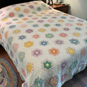 May include: A white bed quilt featuring a repeating pattern of multi-colored floral motifs. Each flower is composed of six-sided hexagonal shapes in shades of yellow, pink, blue, green, and purple, stitched onto a textured white fabric base.