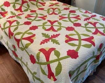 May include: A white quilt with a floral pattern in shades of red, green, and yellow. The quilt is on a bed with a wooden headboard.