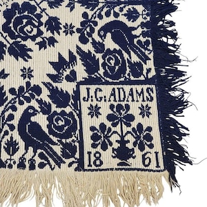 May include: A close-up of a cream-colored textile with a navy blue floral and bird pattern. The textile has a fringed edge and a rectangular label with the text "J.G. ADAMS" and the numbers "1861".