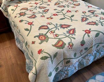 May include: A floral quilt with a cream background and a repeating pattern of red, green, and yellow flowers and leaves. The quilt is on a bed with a wooden headboard. The bed skirt has a blue and green floral pattern. A round rug is visible.