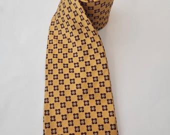May include: A rolled-up gold-colored tie with a repeating pattern of dark brown floral squares. The tie is made of a smooth fabric and is displayed against a white background. The tie is a classic accessory.
