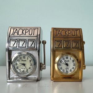 May include: Two slot machine-shaped quartz clocks. One is silver-toned, the other gold-toned. Both have "JACKPOT" at the top and "777" below. Each clock face displays the time and the brand name "Xanadu".