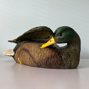 May include: A mallard duck decoy figurine featuring a textured brown body, a dark green head, and a bright yellow beak. The duck is depicted in a resting pose with its head turned toward its back, showcasing detailed feather carvings and a white tail.