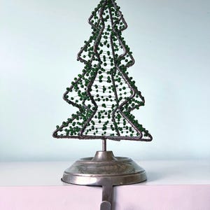May include: A decorative Christmas tree stocking holder featuring a wire frame structure adorned with small green beads. The tree is mounted on a circular metallic base with a downward-facing hook designed to hang from a shelf or mantelpiece.