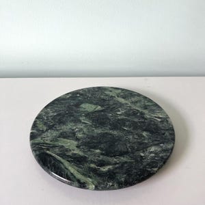 May include: A circular slab of polished dark green marble featuring intricate natural veining in shades of black, forest green, and light gray. The stone surface is smooth and reflective, showcasing a complex, organic pattern throughout the entire piece.