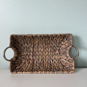 May include: A rectangular woven tray made of brown natural fibers featuring a thick braided pattern throughout the base and sides. The tray includes two circular silver-toned metal handles attached to the left and right sides of the frame.
