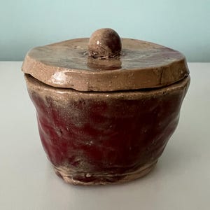 May include: A handmade ceramic jar with a lid. The jar is a deep burgundy color with a textured surface. The lid is a lighter brown with a small, round knob on top. The jar is likely used for storage.