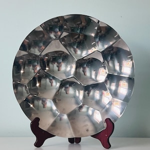 May include: A circular, polished silver-toned metal decorative plate featuring a faceted, geometric surface pattern that creates multiple reflections. The plate is displayed upright on a dark reddish-brown wooden stand with three curved supports.