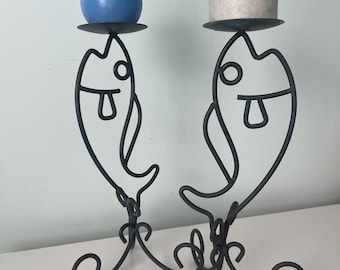 May include: A pair of black metal candle holders shaped like stylized fish, each featuring a circular platform at the top. One holder supports a round blue candle, while the other holds a cylindrical off-white candle. The base has curved wire legs.