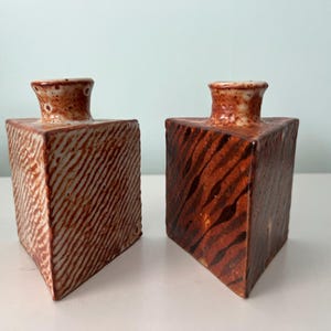May include: Two square ceramic vases with textured surfaces and a reddish-brown color scheme. The vases have a narrow neck and a wide base. One vase has diagonal lines, and the other has a striped pattern.