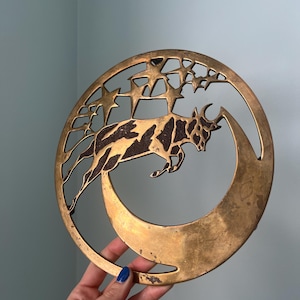 May include: A circular brass wall hanging featuring a cutout design of a bull leaping over a crescent moon, with several stars positioned above the bull. The metal has a weathered, golden-brown finish with dark, textured detailing on the bull.