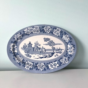 May include: An oval ceramic platter featuring a blue and white transferware pattern. The center depicts a landscape with pagodas, trees, and a bridge. The wide rim is decorated with a dense floral border of blossoms and leaves in blue and white.