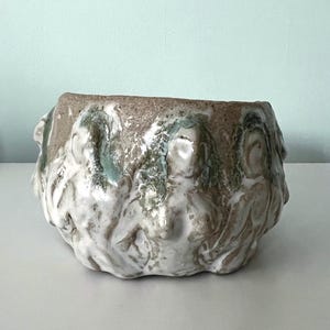 May include: A handcrafted ceramic bowl with a textured surface. The bowl features a light brown rim and is adorned with sculpted figures in white and green. The bowl is a unique piece of art.