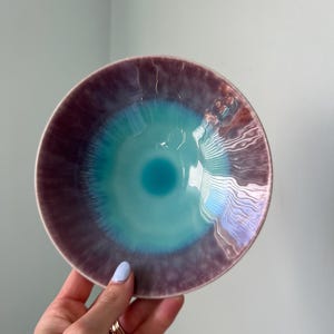 May include: A round ceramic bowl featuring a vibrant gradient glaze that transitions from a deep, saturated turquoise center to a soft, muted purple rim. The surface has a glossy, reflective finish with subtle, fine crackle textures throughout.