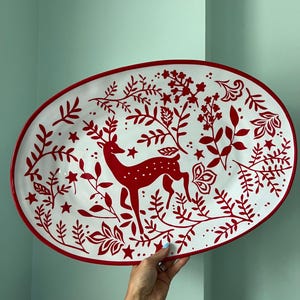 May include: An oval-shaped serving platter featuring a white base with a vibrant red pattern. The design depicts a central deer surrounded by intricate botanical elements, including leafy branches, small flowers, and scattered stars.