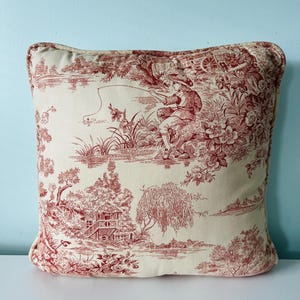 May include: A square throw pillow featuring a cream-colored fabric with a detailed red toile de jouy print. The pattern depicts a pastoral scene with a person fishing by a river, lush trees, floral elements, and a small cottage structure.